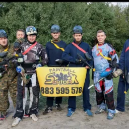 Grupa osób w strojach do paintballa, trzymających markery i żółty baner z napisem 'Paintball Sektor' i numerem telefonu, na tle drzew.