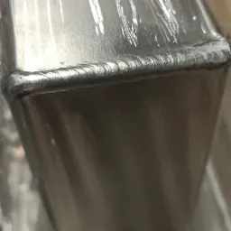 Szlifowany profil zamknięty ze stali nierdzewnej, z widocznymi śladami spawania, owinięty folią ochronną, zamontowany na metalowej podstawie z otworami.