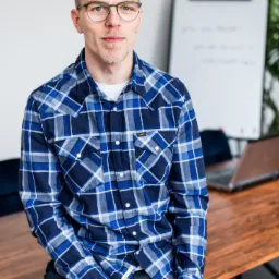 Młody mężczyzna w okularach i niebieskiej koszuli w kratę siedzi na drewnianym biurku, trzymając marker, z tablicą suchościeralną i laptopem w tle.