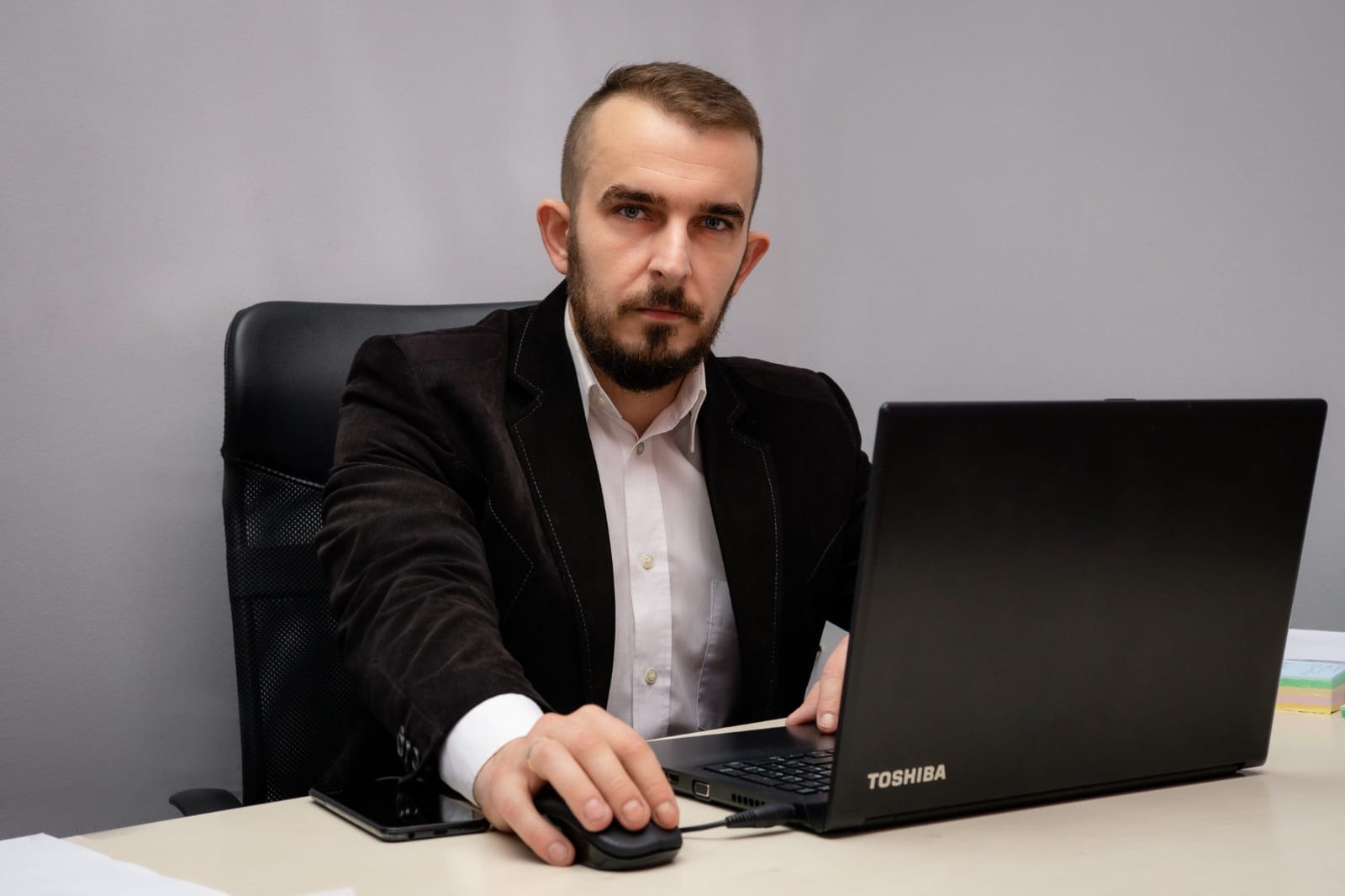 Mężczyzna z brodą w ciemnej marynarce i białej koszuli siedzi przy biurku z laptopem Toshiba i myszką, spoglądając wprost.
