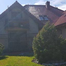 Dom jednorodzinny w trakcie prac elewacyjnych. Widoczne rusztowania, zabezpieczone okna folią, świeżo nałożony tynk. Czerwona dachówka i zieleń trawnika w słoneczny dzień.