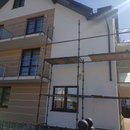 Dwupiętrowy dom z balkonami w trakcie prac elewacyjnych. Widoczne rusztowania, jasna elewacja z beżowymi akcentami. Słoneczny dzień.