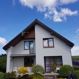 Dwukondygnacyjny dom z jasną elewacją, brązowymi ramami okiennymi i dachem. Widoczne krzewy i dziecko w ogrodzie. Antena na dachu. Błękitne niebo z chmurami w tle.