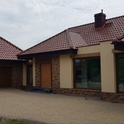 DARBUD DARIUSZ DANILCZYK - Dom jednorodzinny z czerwoną dachówką i elewacją w odcieniach beżu i brązu. Widoczne okna z drewnianymi ramami i ceglana podmurówka. Na podjeździe kosiarka.