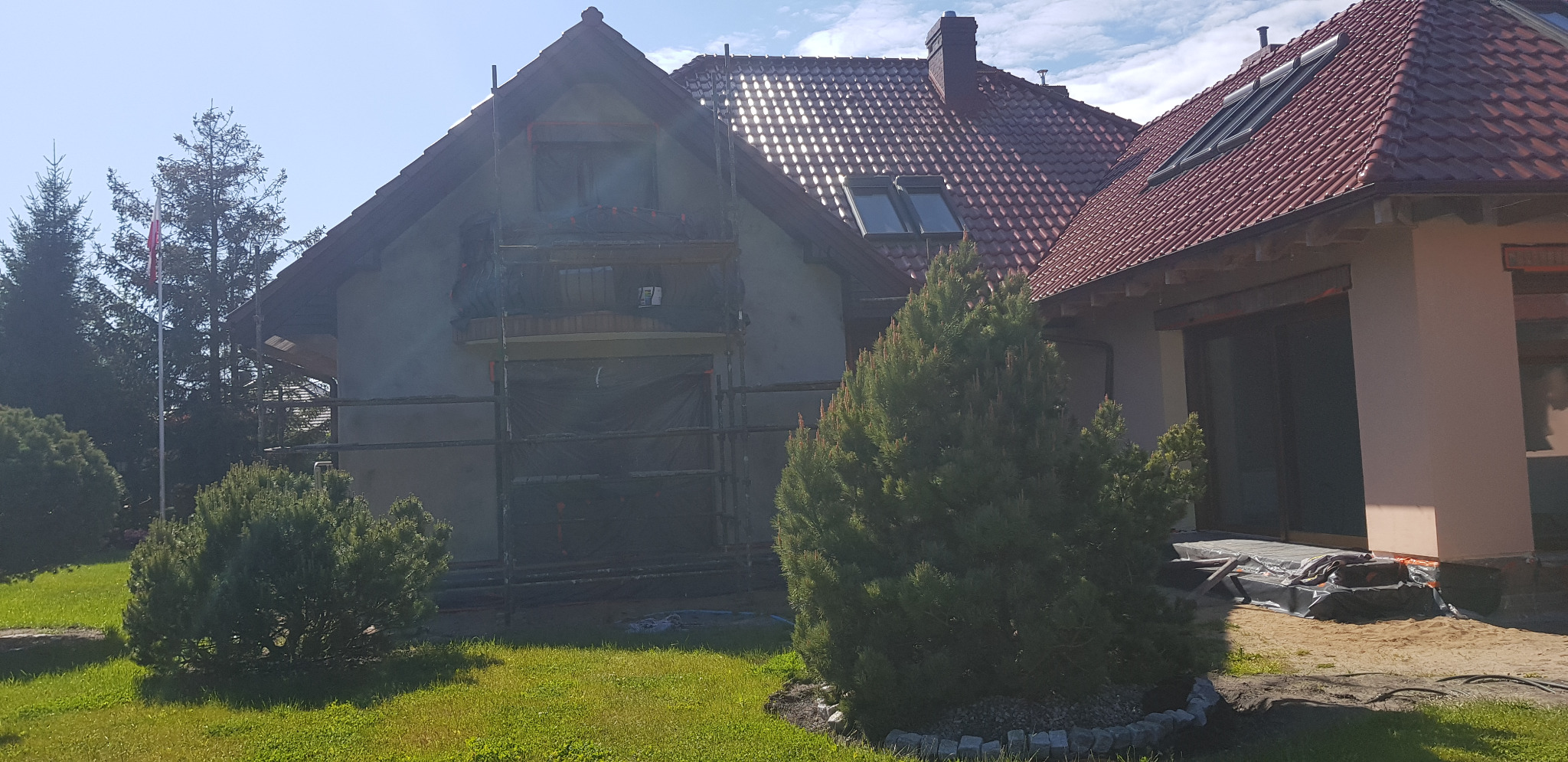 Dom jednorodzinny w trakcie prac elewacyjnych. Widoczne rusztowania, zabezpieczone okna folią, świeżo nałożony tynk. Czerwona dachówka i zieleń trawnika w słoneczny dzień.