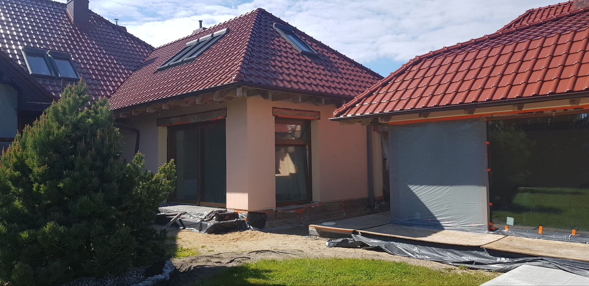 Dom jednorodzinny z nową, jasną elewacją w trakcie wykańczania. Widoczne zabezpieczenia okien folią i przygotowany teren wokół budynku. Czerwona dachówka.