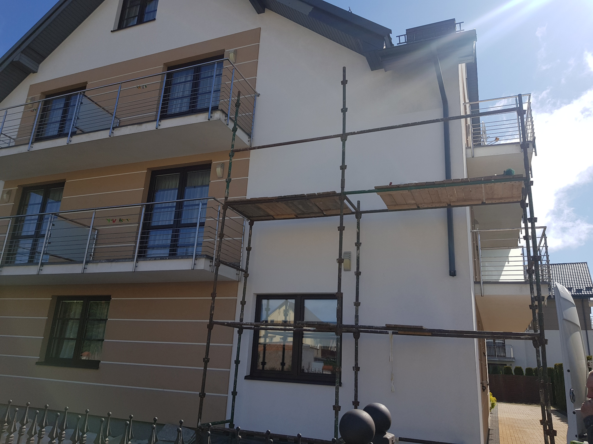 Dwupiętrowy dom z balkonami w trakcie prac elewacyjnych. Widoczne rusztowania, jasna elewacja z beżowymi akcentami. Słoneczny dzień.