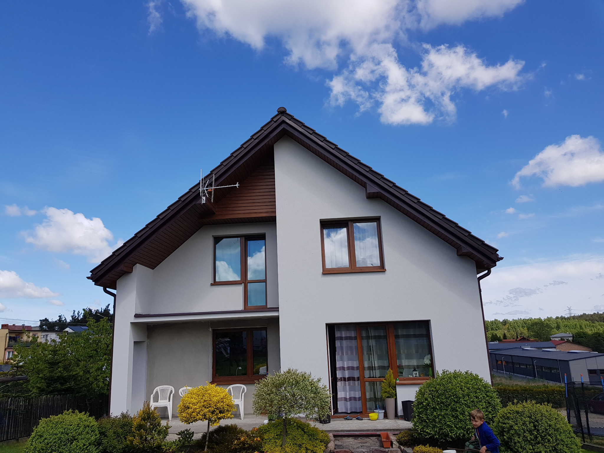 Dwukondygnacyjny dom z jasną elewacją, brązowymi ramami okiennymi i dachem. Widoczne krzewy i dziecko w ogrodzie. Antena na dachu. Błękitne niebo z chmurami w tle.