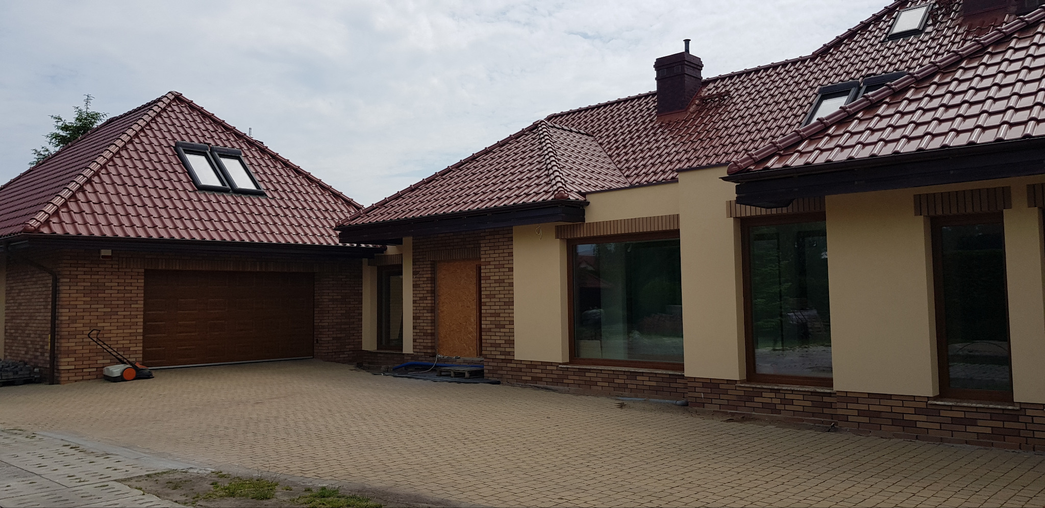 Dom jednorodzinny z czerwoną dachówką i elewacją w odcieniach beżu i brązu. Widoczne okna z drewnianymi ramami i ceglana podmurówka. Na podjeździe kosiarka.