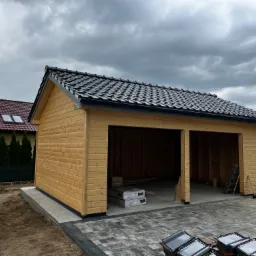 Nowo wybudowany, dwustanowiskowy garaż z drewna, z czarną dach&oacute;wką, na posesji z kostką brukową, widok z zewnątrz, pochmurne niebo.