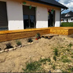 Nowoczesny taras drewniany z desek ułożonych poziomo, przylegający do białego domu z szarymi roletami, z widoczną paletą i młodymi roślinami w ziemi.