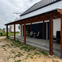 Piotr Szymczak - Drewniana konstrukcja pergoli przylegająca do domu z ciemnymi elementami, na tarasie stolik z krzesłami, w tle budowa domu w trakcie realizacji.