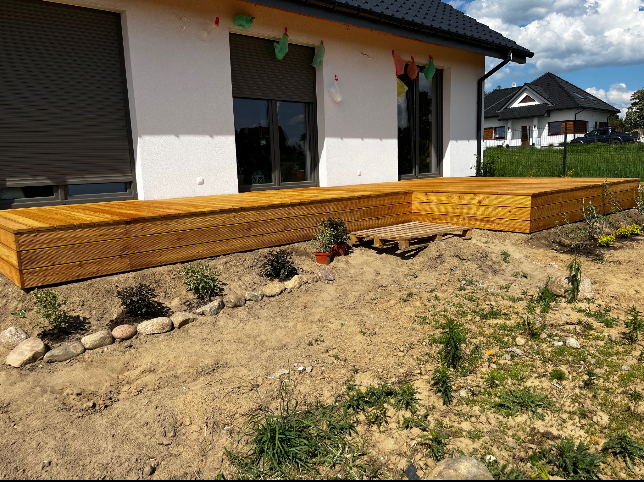 Nowoczesny taras drewniany z desek ułożonych poziomo, przylegający do białego domu z szarymi roletami, z widoczną paletą i młodymi roślinami w ziemi.