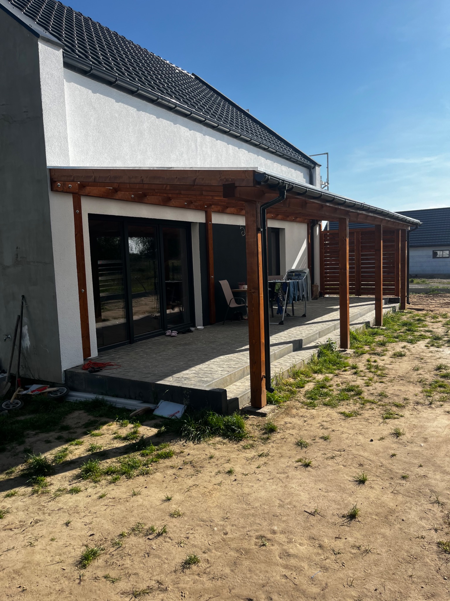 Drewniana konstrukcja zadaszenia przylegająca do budynku z szarymi płytkami tarasowymi i widocznym praniem na suszarce, na tle błękitnego nieba.