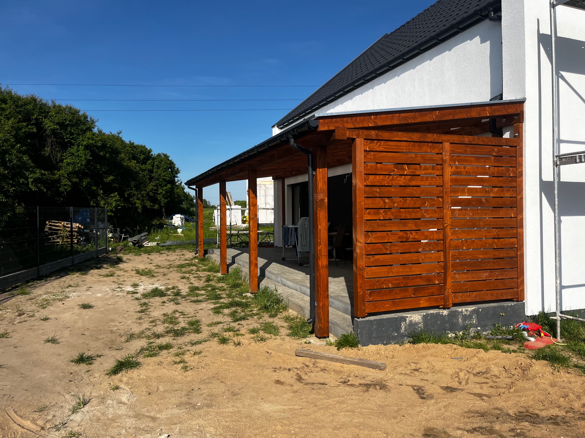 Drewniana konstrukcja z poziomych desek, prawdopodobnie zadaszenie tarasu lub pergola, przylegająca do białego domu z czarnym dachem, widoczna ziemia i trawa, w tle drzewa.