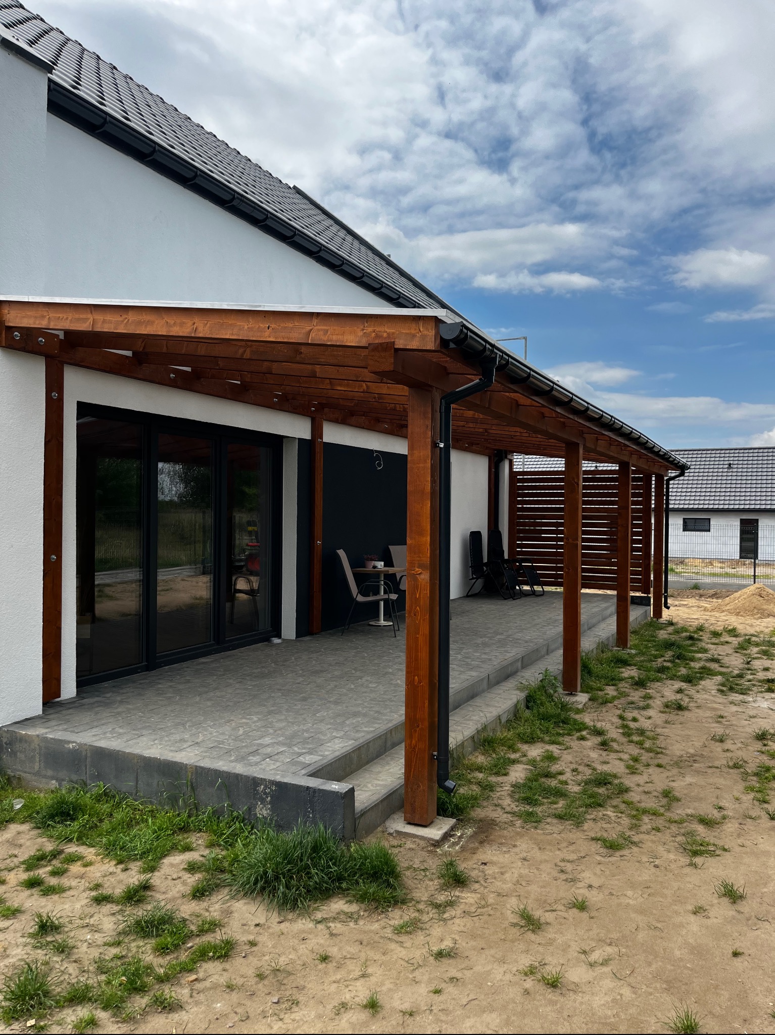 Drewniana pergola przy nowoczesnym domu z przesuwnymi drzwiami, szara kostka brukowa na tarasie, widoczne krzesła i stolik. Na drugim planie budynek w trakcie budowy i błękitne niebo z chmurami.