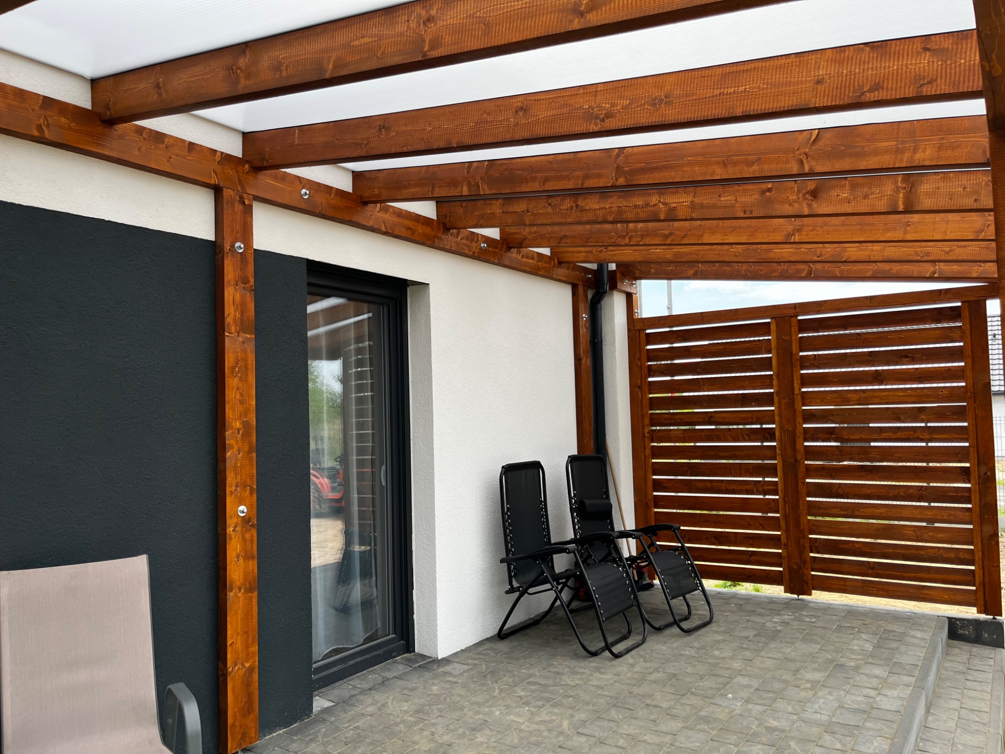Drewniana pergola z przezroczystym zadaszeniem, przylegająca do ściany budynku pomalowanej na biało i grafitowo, z trzema czarnymi leżakami na szarym brukowanym tarasie.