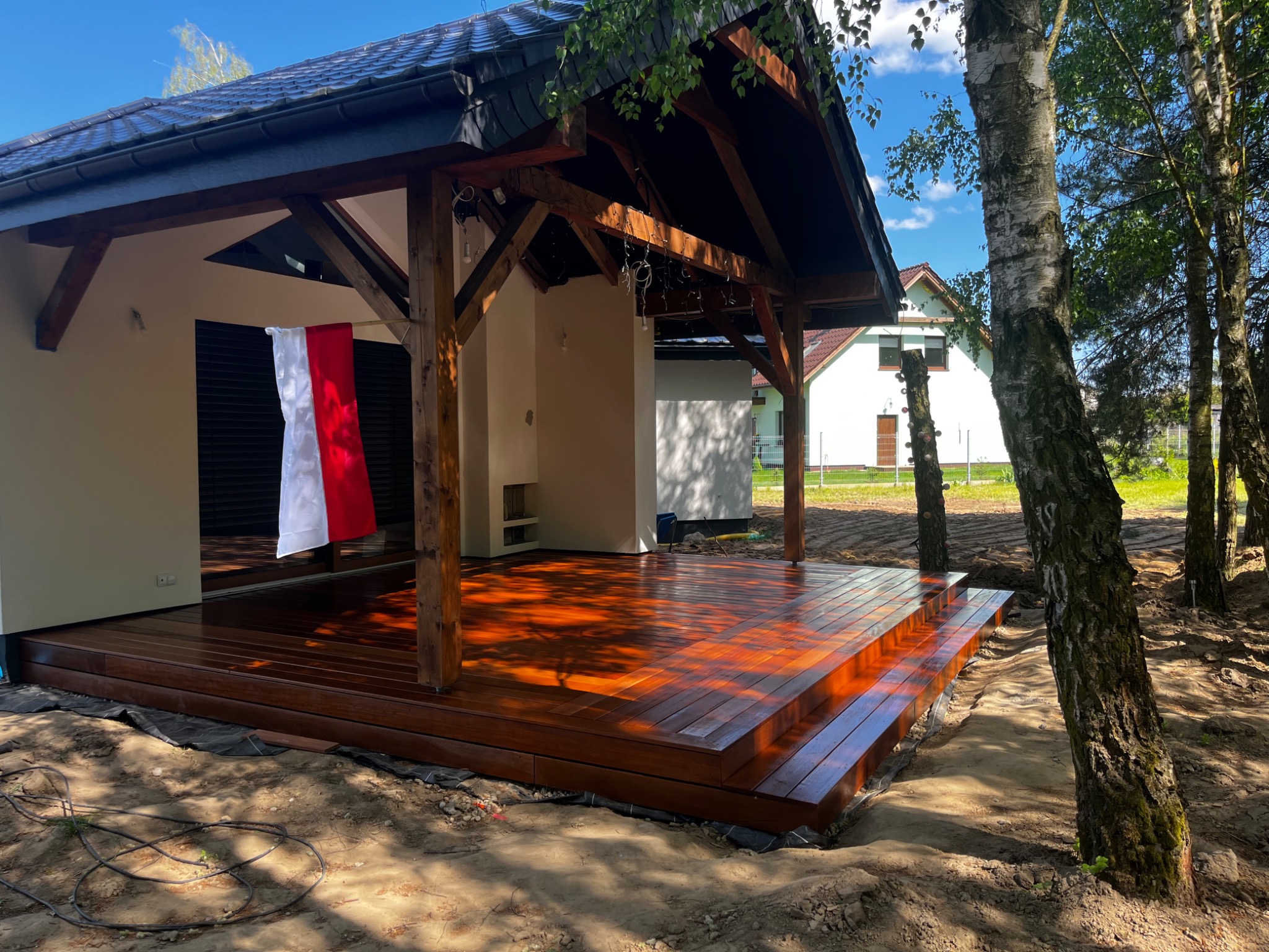 Zadaszony taras z drewnianą podłogą, polakierowany na wysoki połysk, z widoczną biało-czerwoną flagą, w otoczeniu drzew i niezabudowanej przestrzeni.