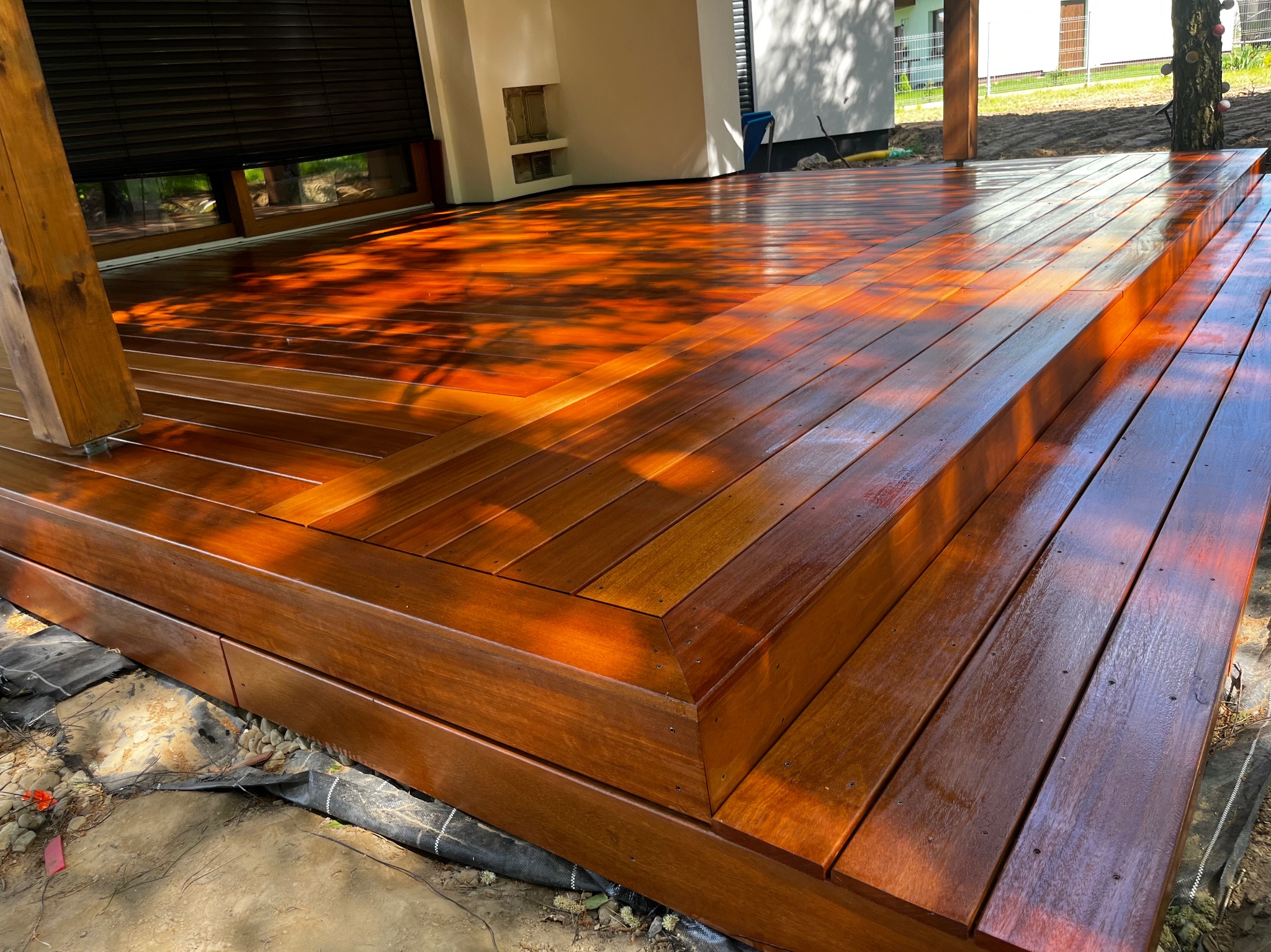 Świeżo polakierowany, drewniany taras z dwoma stopniami w Dobrzanach, z widocznymi refleksami światła na powierzchni desek i czarną geowłókniną u podstawy konstrukcji.