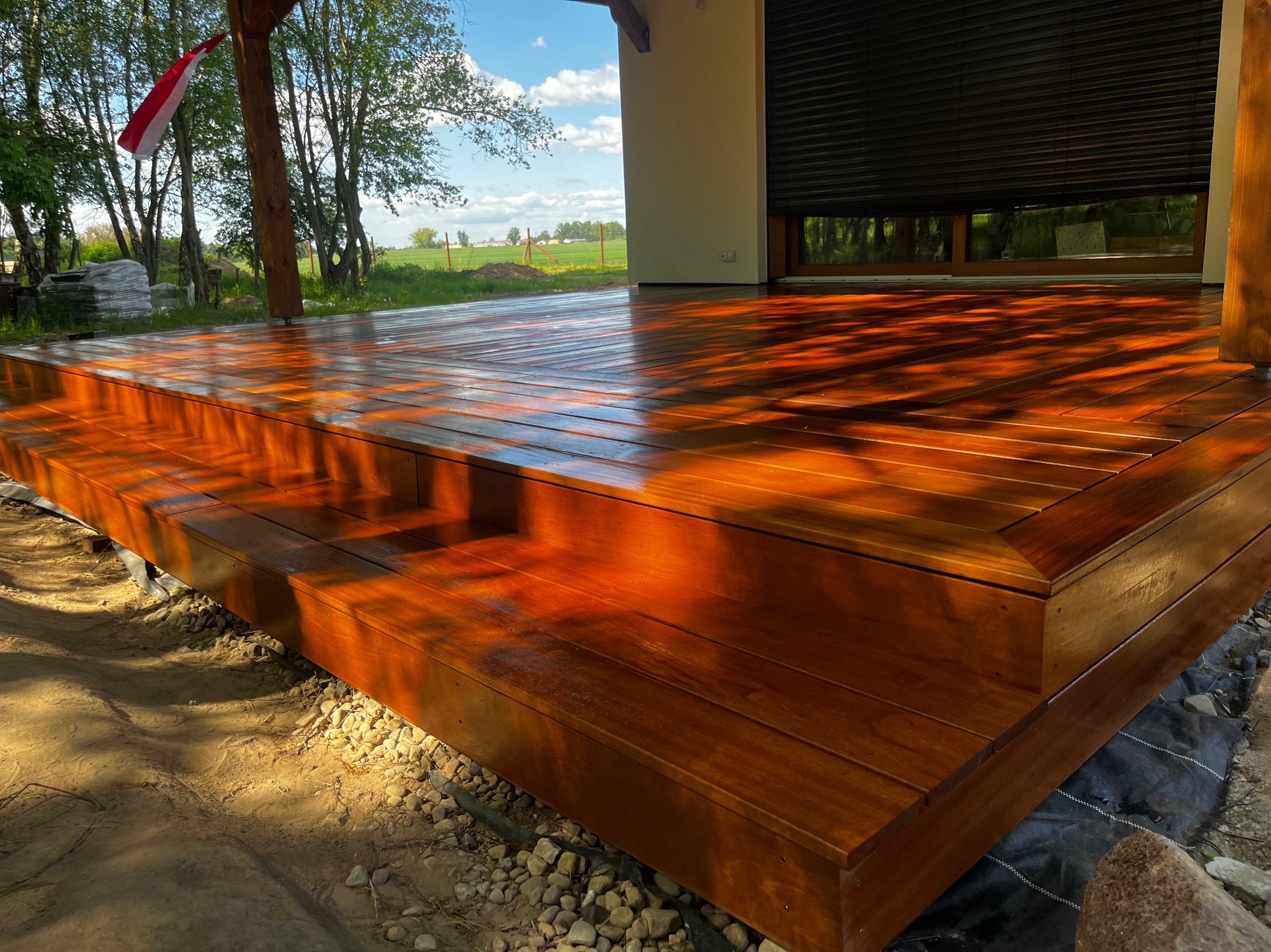 Błyszczący, świeżo polakierowany taras drewniany z niskimi schodkami, widok na zielony krajobraz i dom z żaluzjami, w tle sterta materiałów budowlanych.