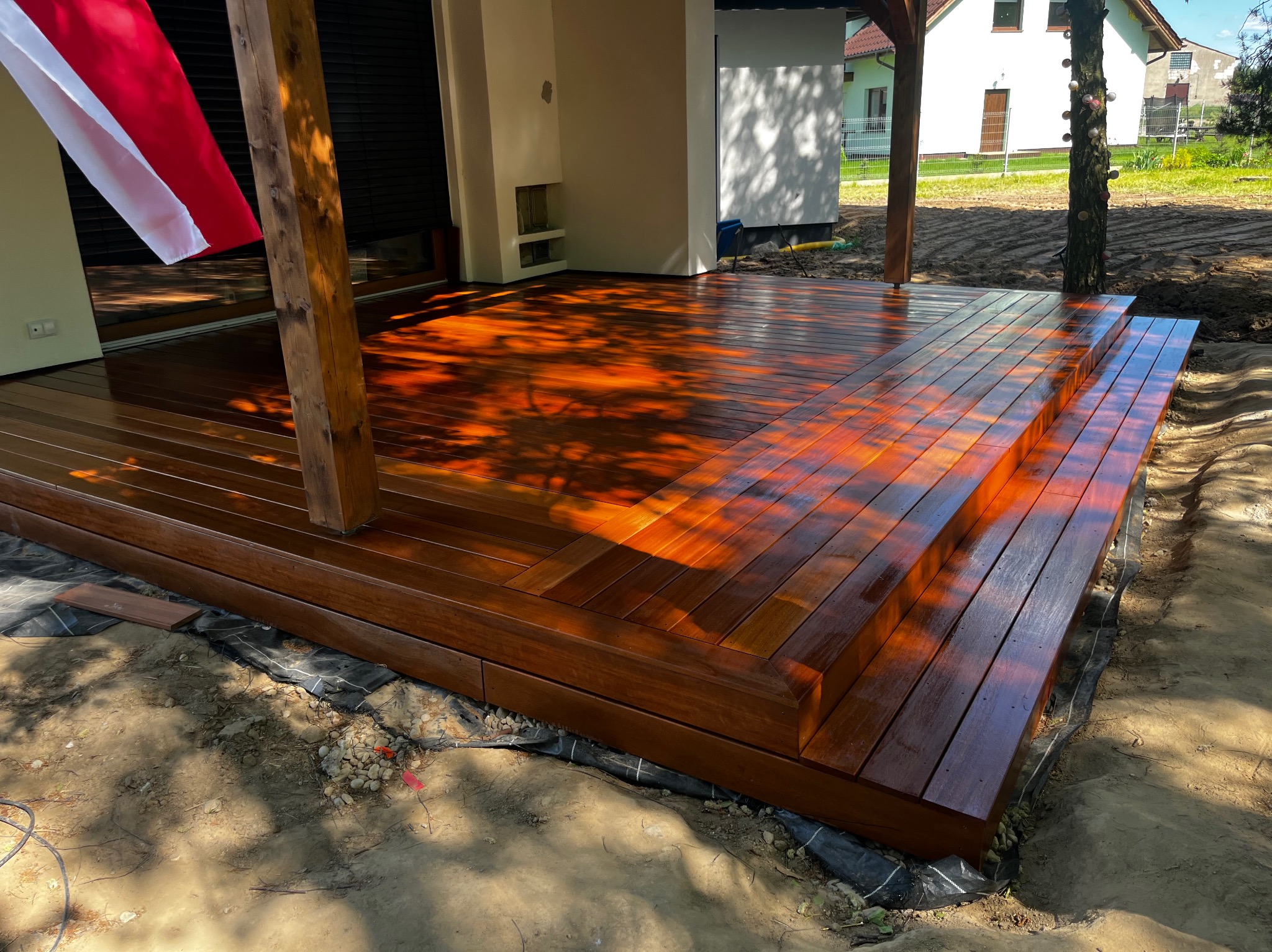 Świeżo polakierowany taras z drewna egzotycznego z trzema stopniami, widoczne refleksy światła, w tle dom z białymi ścianami i fragment polskiej flagi.