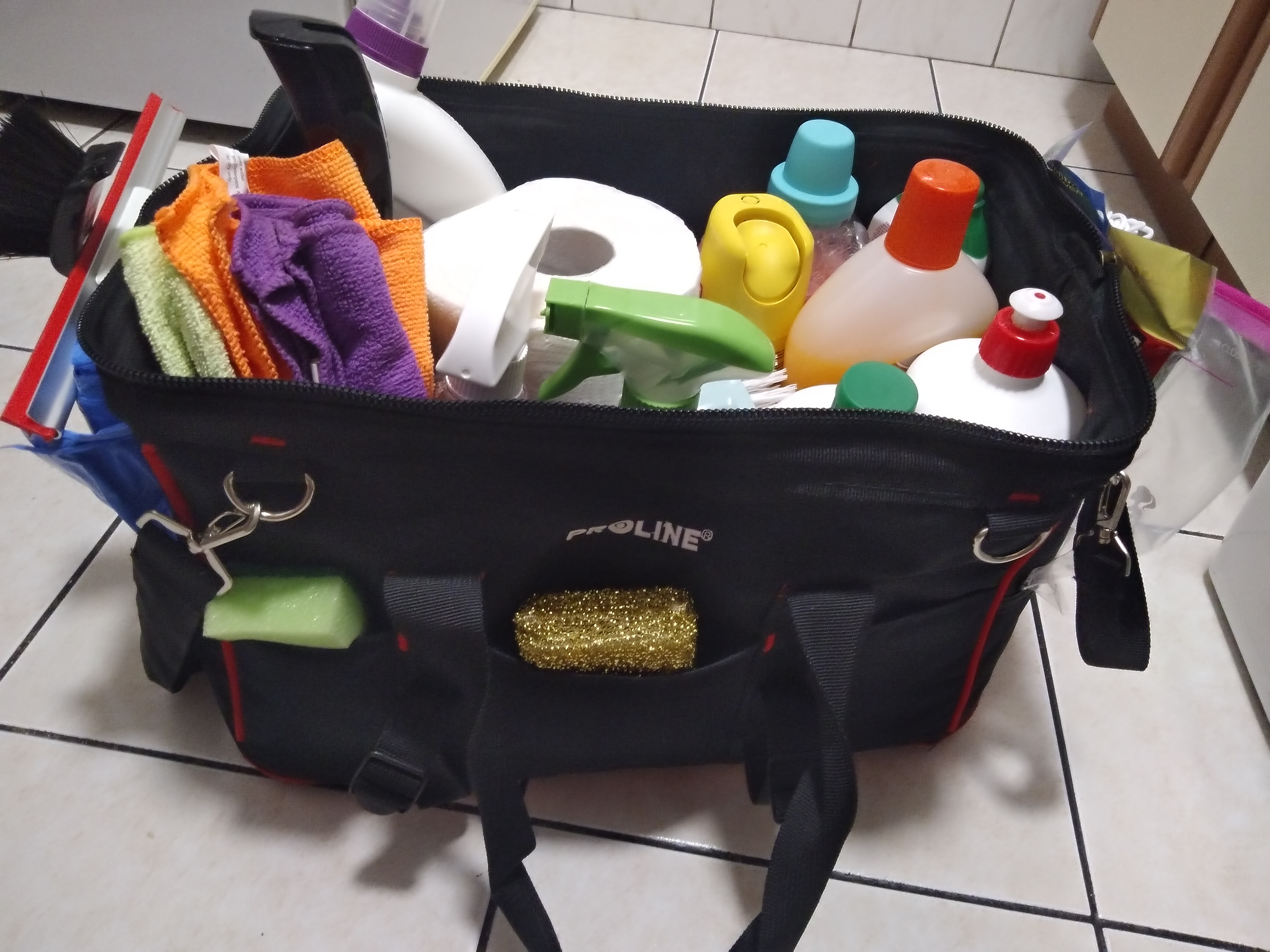 Czarna torba narzędziowa marki Proline wypełniona różnorodnymi środkami czystości, ściereczkami z mikrofibry w odcieniach zieleni, pomarańczy i fioletu, spryskiwaczem, rolką papieru oraz gąbkami...