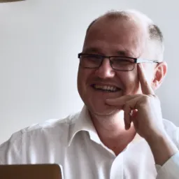 Uśmiechnięty mężczyzna w okularach, oparty palcem o skroń, częściowo widoczny laptop, lampa sufitowa w tle.