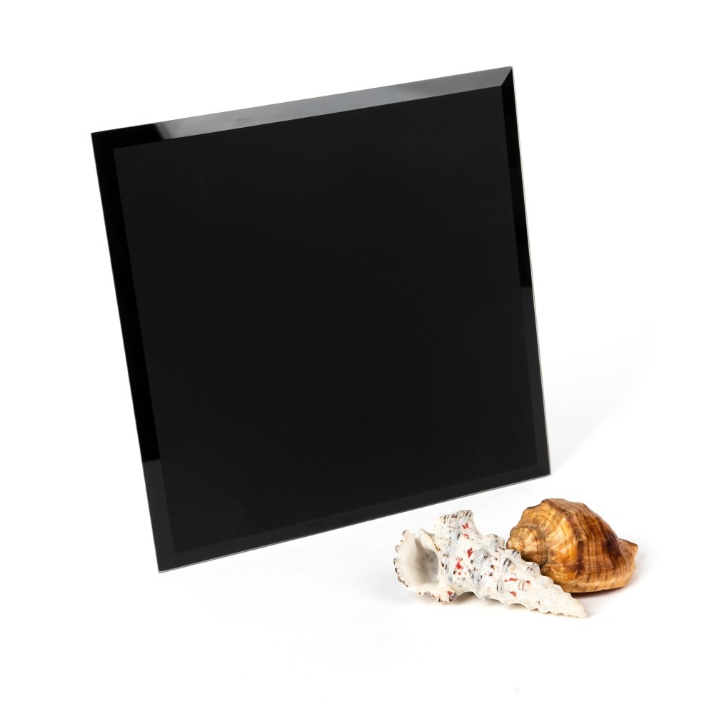Czarne lustro kwadratowe z fazowanymi krawędziami, ustawione ukośnie na białym tle obok dwóch muszli.