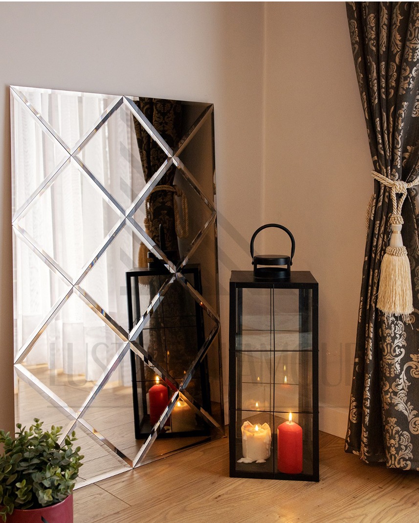 Dekoracyjne lustro ścienne z fazowanymi rombami, odbijające płomień świecy w lampionie, obok zasłona z ciemnego wzorzystego materiału i doniczka z rośliną.