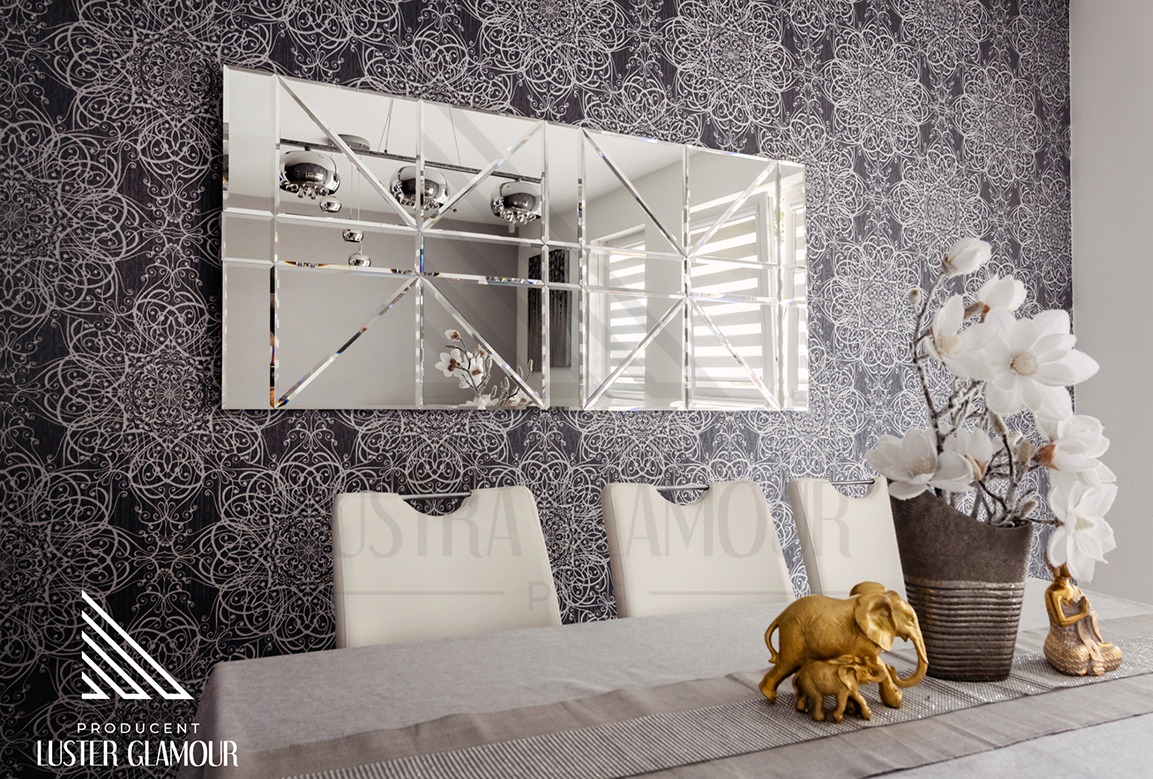 Prostokątne lustro złożone z kwadratowych elementów z fazowanymi krawędziami na ścianie z wzorzystą tapetą, odbijające lampę i okno, stół z dekoracjami: figurki słoni i kwiaty.