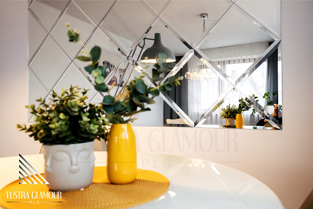 Dekoracyjne lustro ścienne z fazowanych rombów, odbijające fragment pokoju z lampą i zasłonami, na pierwszym planie biały stół z żółtą podkładką i dwoma wazonami z roślinami, widoczne logo firmy...