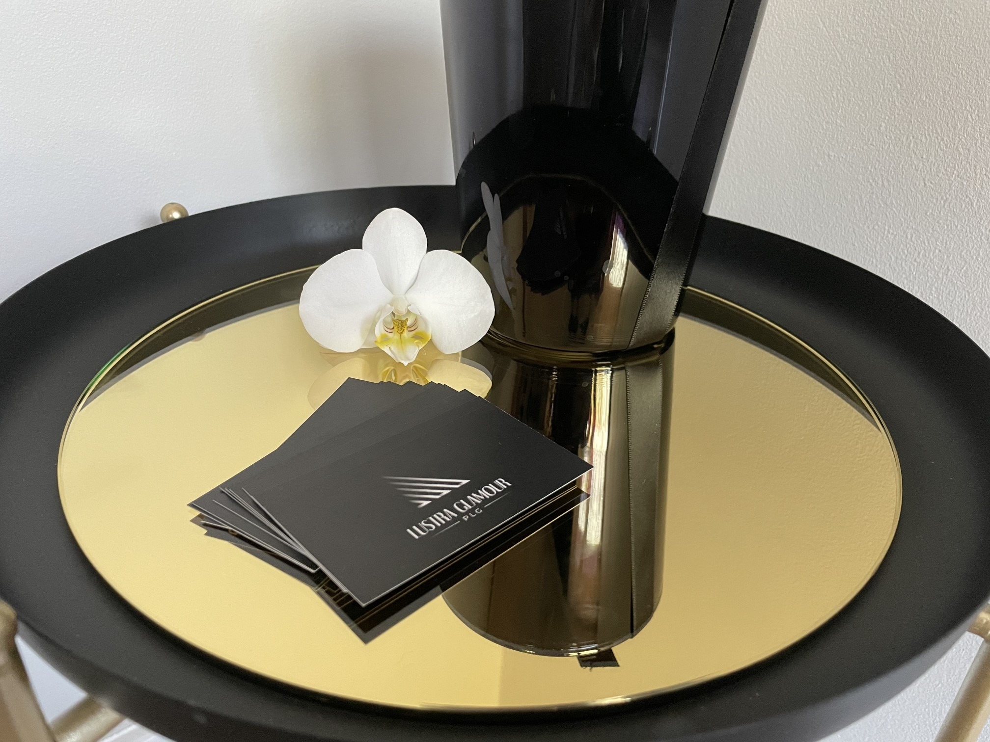Wizytówki firmy Lustra Glamour PLC leżące na złotym, okrągłym lustrze, obok białego storczyka i czarnego wazonu.