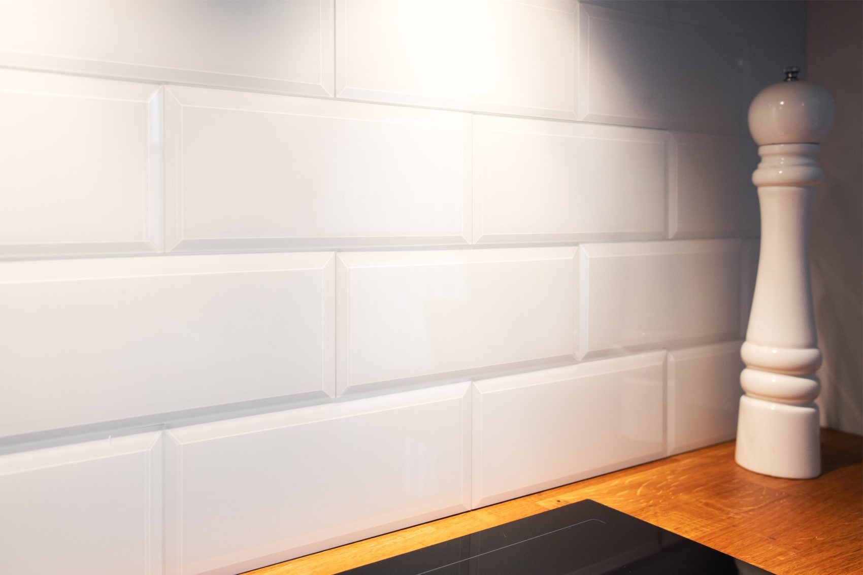 Białe, prostokątne płytki ścienne, ułożone w rzędach, z widoczną drewnianą powierzchnią blatu i fragmentem płyty indukcyjnej oraz ceramicznym młynkiem do pieprzu.