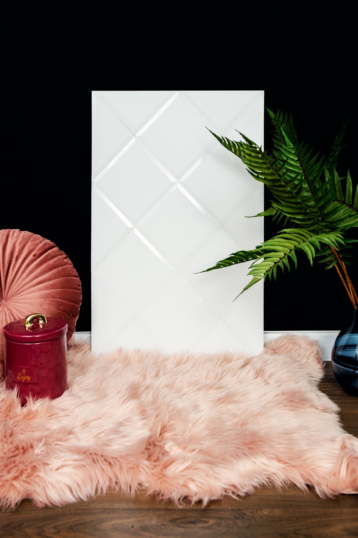 Dekoracyjny panel ścienny z białego szkła z romboidalnym wzorem, ustawiony na puszystym, różowym dywaniku, obok poduszki i wazonu z paprotką na drewnianej podłodze.