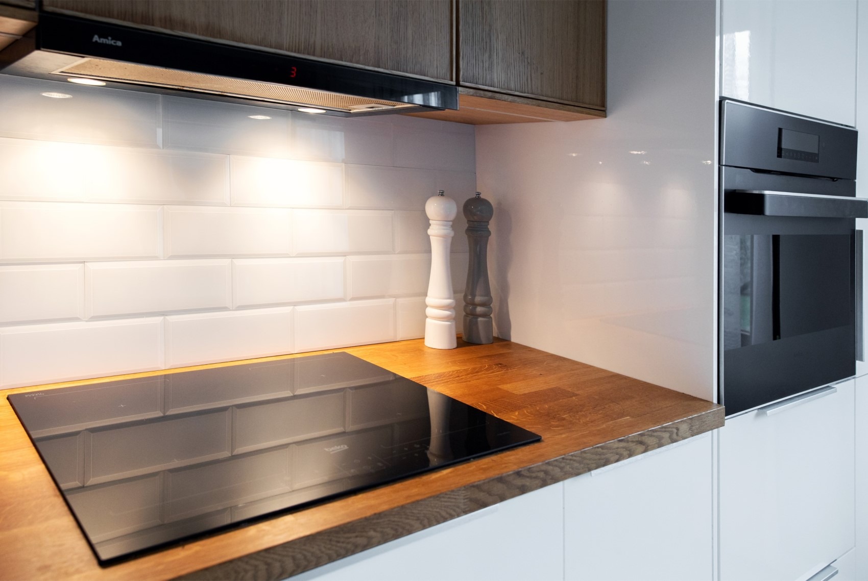 Fragment nowoczesnej kuchni z drewnianym blatem, płytą indukcyjną Beko, białymi kaflami w stylu retro, okapem Amica i białymi szafkami z piekarnikiem w zabudowie.