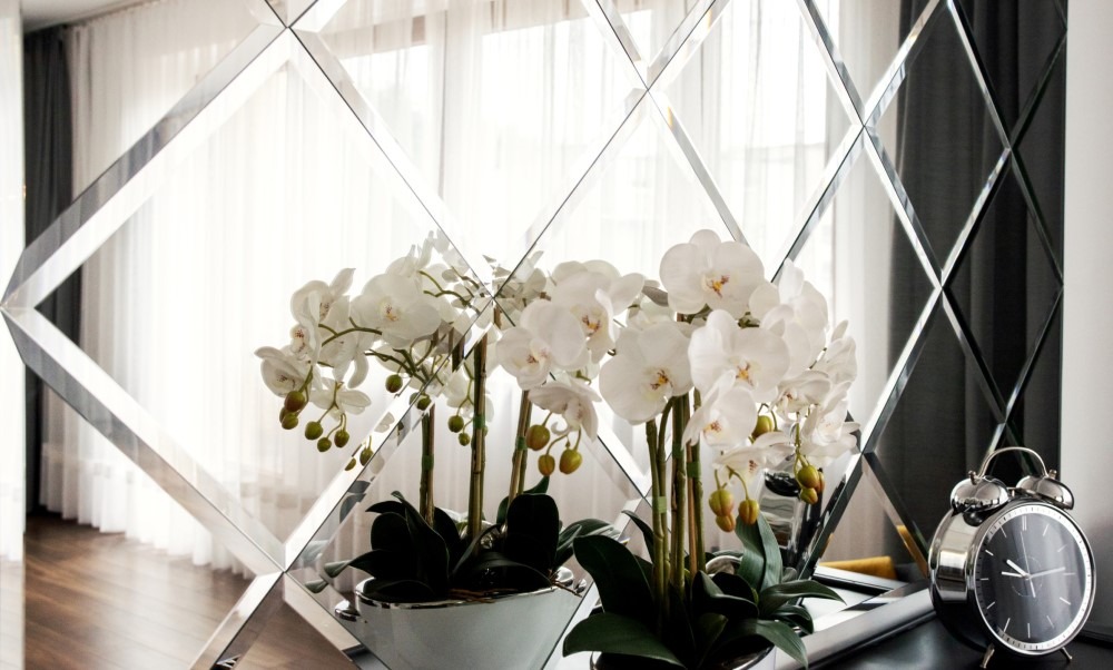 Dekoracyjna ściana z lustrzanych rombów odbijająca światło, z białymi orchideami w srebrnych donicach i srebrnym budzikiem na czarnym blacie.