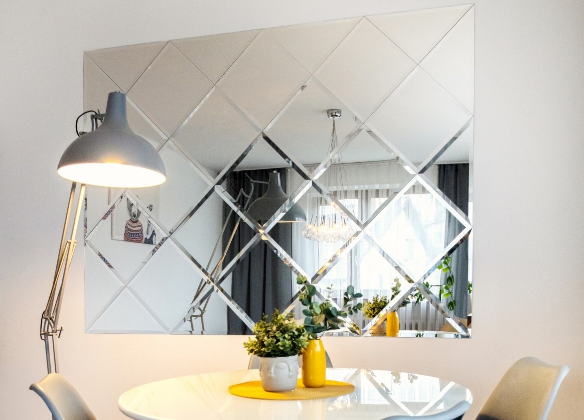 Ściana z dekoracyjnych, lustrzanych kwadratów w jadalni, odbijająca lampę i okno, z widocznym stołem i roślinami.