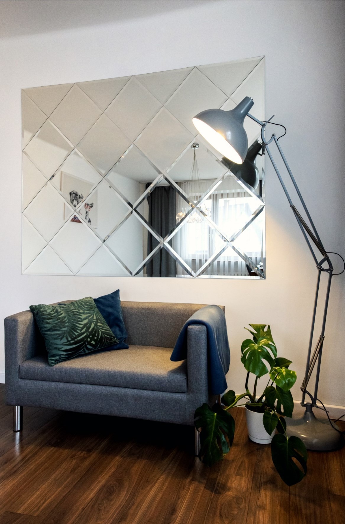 Ściana z lustrzanych płytek ułożonych w romby nad szarą sofą z zieloną i niebieską poduszką, obok lampa podłogowa i roślina w doniczce.