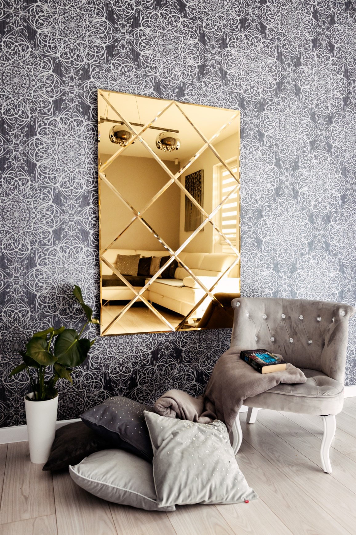 Złote lustro ścienne z kwadratowych tafli, odbijające wnętrze salonu, ustawione na tle wzorzystej tapety, obok szarego fotela z książką i poduszkami na podłodze.