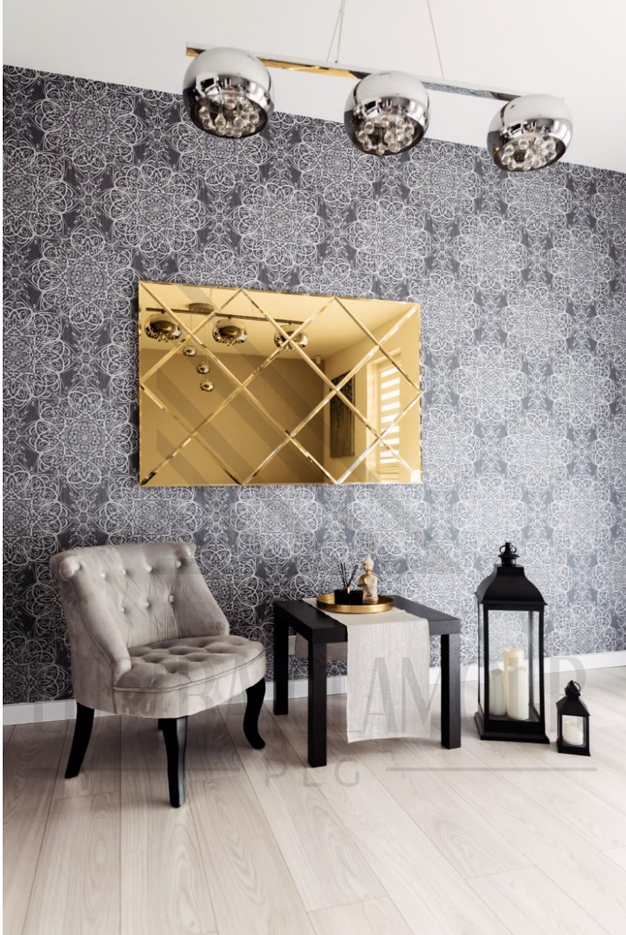 Złote lustro z fazowanymi krawędziami w kształcie rombów, zawieszone na wzorzystej szarej ścianie, odbija nowoczesne lampy sufitowe. Na pierwszym planie szary, tapicerowany fotel i czarny stolik...