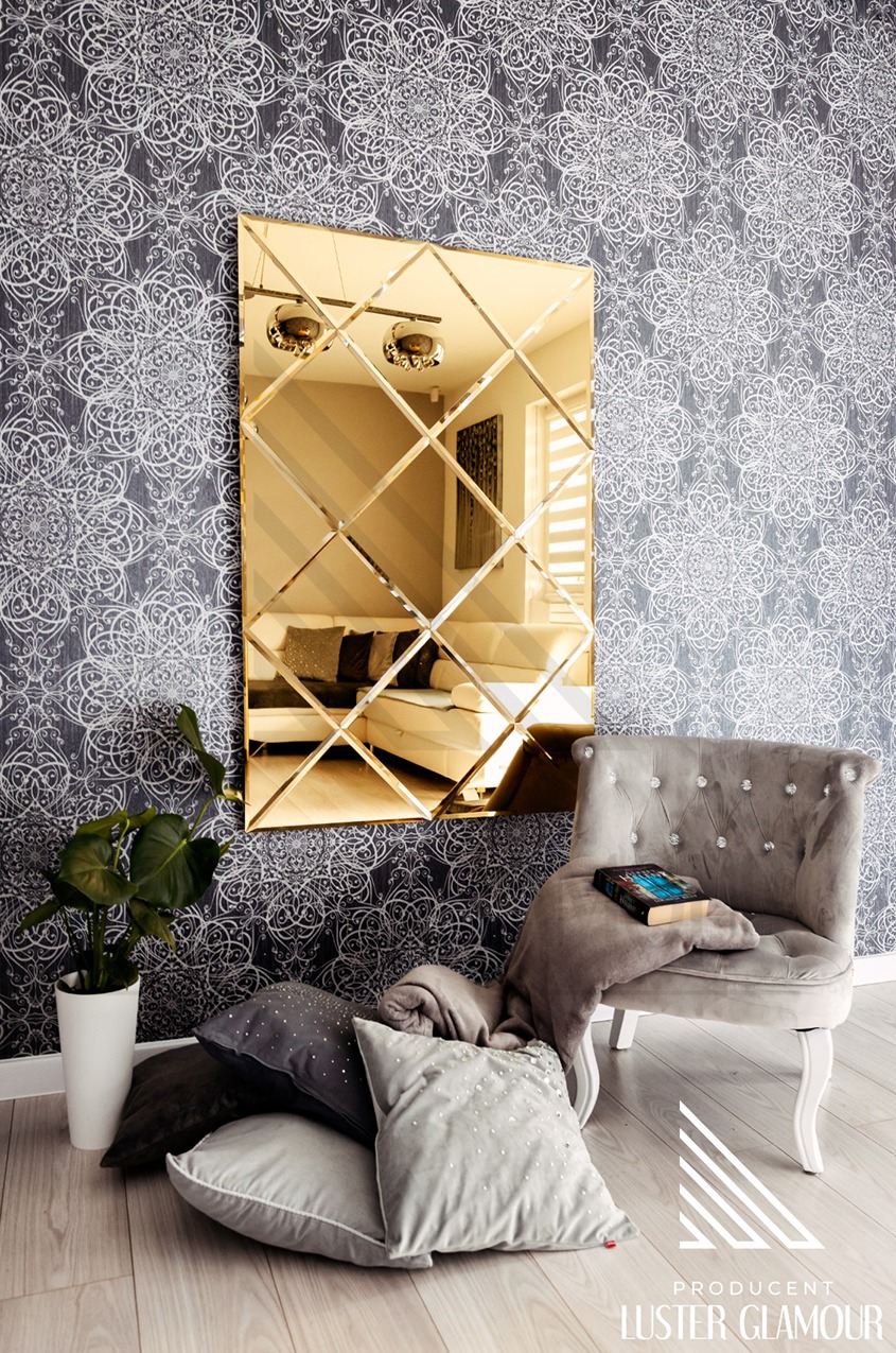 Złote lustro złożone z kwadratowych elementów, umieszczone na ścianie z wzorzystą tapetą, odbija wnętrze pokoju z sofą; obok lustra stoi szary fotel z poduszkami i pledem, a na podłodze leżą...