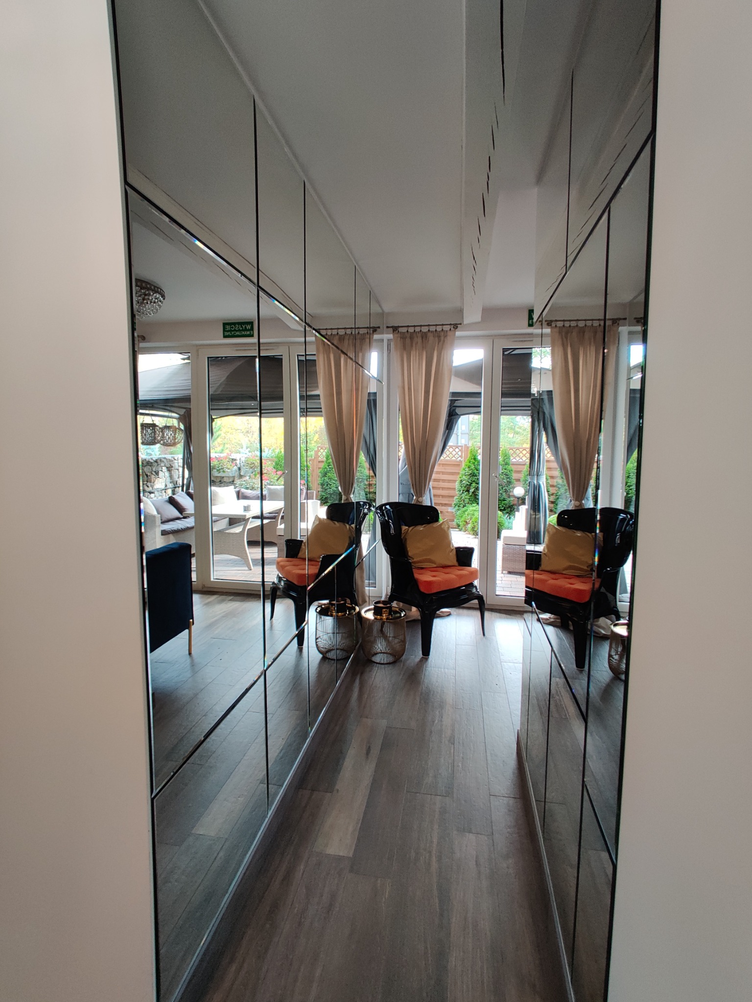 Ściana z pionowych luster w wąskim korytarzu, odbicie dwóch czarnych foteli z pomarańczowymi poduszkami, widok na taras z meblami ogrodowymi przez przeszklone drzwi.