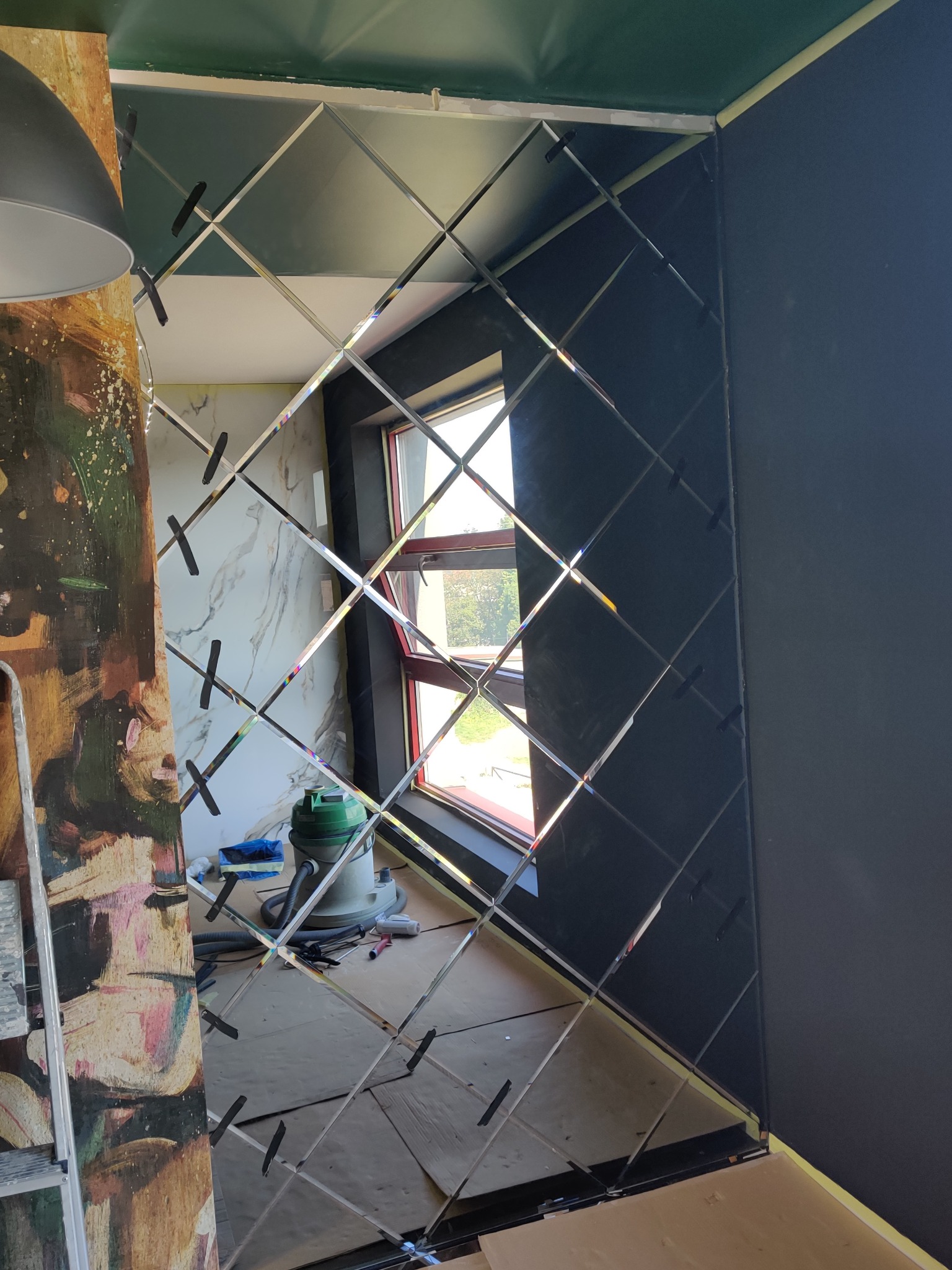 Montaż dekoracyjnych luster w kształcie rombów na ścianie, widoczne przyssawki mocujące, obok drabina malarska i odkurzacz.