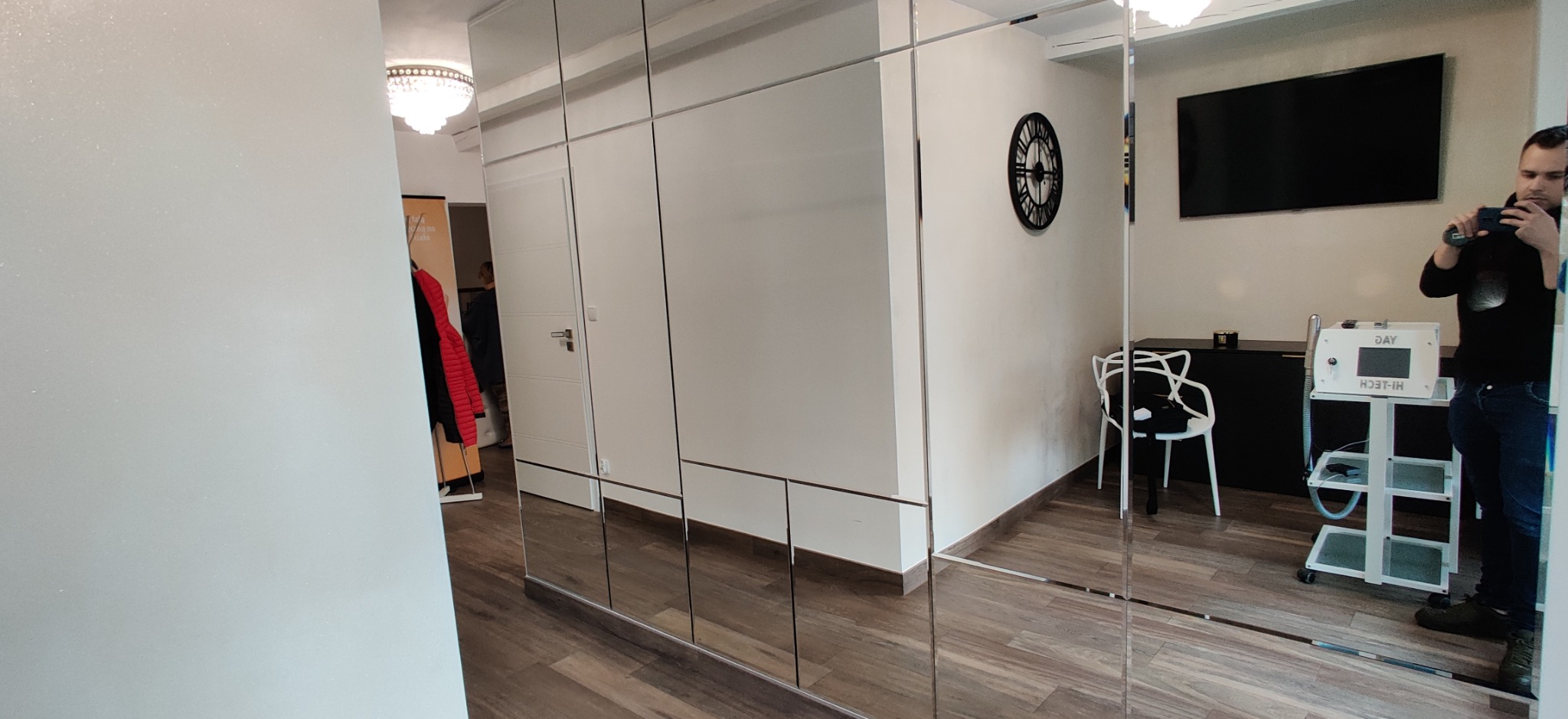 Wielkopowierzchniowa szafa z lustrzanymi drzwiami przesuwnymi, odbijająca wnętrze pokoju z widocznym zegarem ściennym, telewizorem i osobą robiącą zdjęcie telefonem.