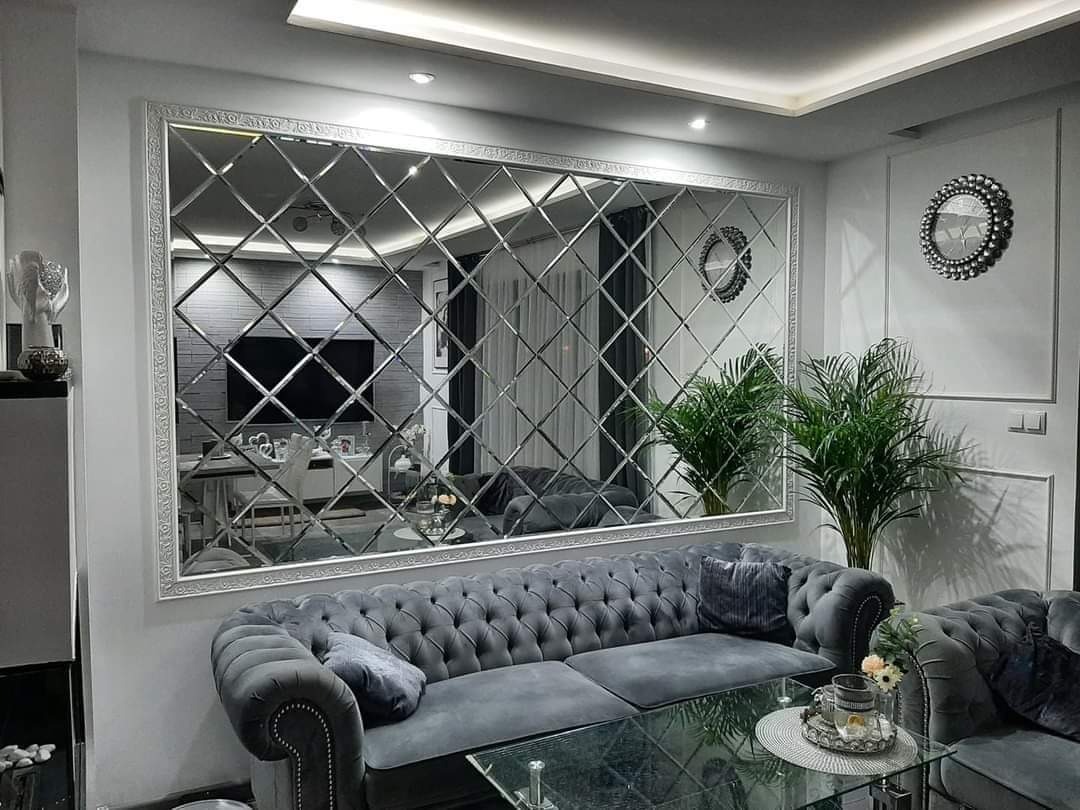 Duże lustro ścienne z fazowanych kwadratów w ozdobnej, srebrnej ramie, odbijające wnętrze salonu z szarą sofą chesterfield i szklanym stolikiem kawowym.