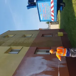 Pracownik w pomarańczowej kamizelce i kasku myje elewację dwukolorowego budynku za pomocą myjki ciśnieniowej, widoczny efekt czyszczenia oraz podnośnik z reklamą firmy.