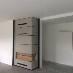 Wnętrze w trakcie prac wykończeniowych: zabudowa kominkowa z betonu architektonicznego z czarnymi akcentami, ściany pomalowane na biało, widoczne przygotowania do dalszych prac w sąsiednim...
