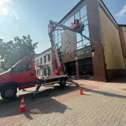 Czerwony podnośnik koszowy czyści szklaną fasadę budynku Eldorado, z widocznymi odbiciami, na tle błękitnego nieba. Pomarańczowe pachołki ostrzegawcze na chodniku.