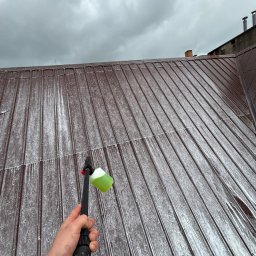 DIZ POL Podnośnik koszowy, Wycinka drzew, Mycie ciśnieniowe - Czyszczenie brązowego dachu z blachy za pomocą myjki ciśnieniowej z zielonym detergentem, widok z dołu na pochmurne niebo.