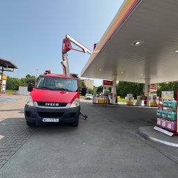 DIZ POL Podnośnik koszowy, Wycinka drzew, Mycie ciśnieniowe - Czerwony samochód Iveco z podnośnikiem koszowym przy stacji benzynowej Shell. Widoczny wysięgnik i elementy konstrukcyjne stacji. Słoneczny dzień.